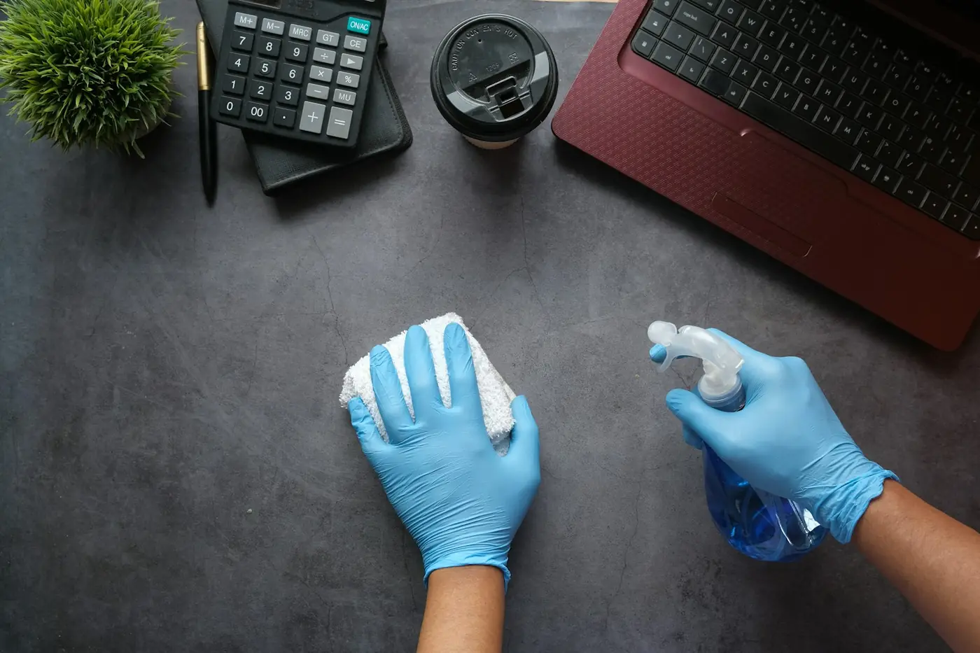 Hände in blauen Handschuhen wischen mit Sprühflasche und Tuch einen dunklen Schreibtisch ab.